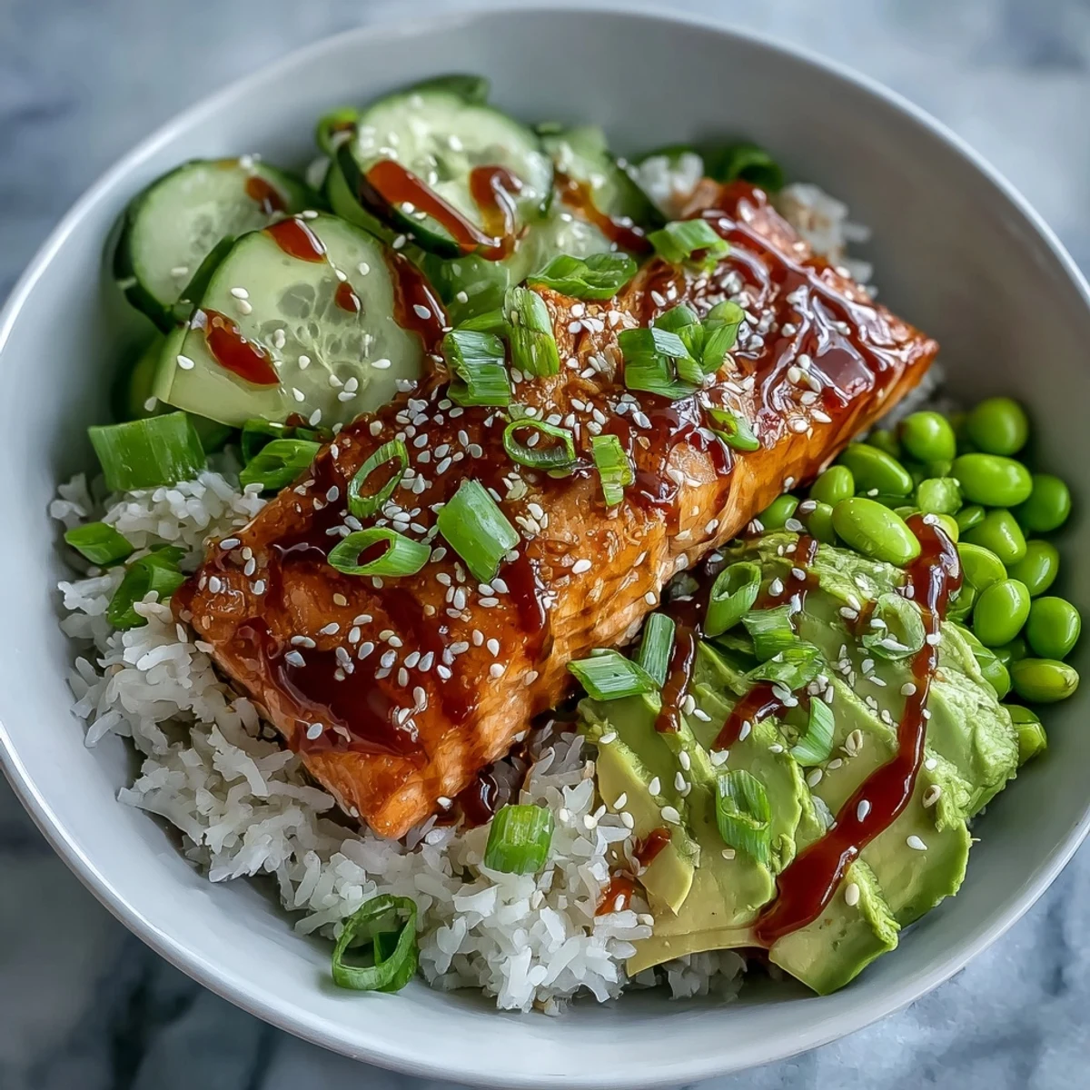 Golden brown salmon fillets glazed with sticky honey sriracha sit atop fluffy jasmine rice in a white bowl. Sliced cucumber, creamy avocado, and bright green edamame add fresh textures alongside a drizzle of spicy mayo.
